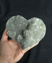 Load image into Gallery viewer, Hand holding a green amethyst crystal rock in the shape of a heart against a black background
