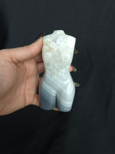 Load image into Gallery viewer, Hand holding a white and blue agate in the shape of a female body against a black background
